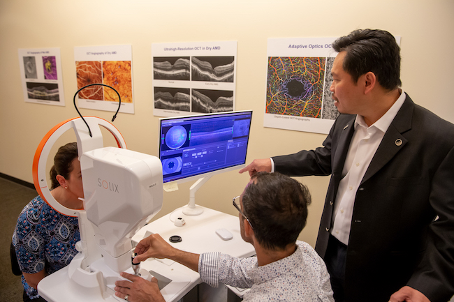 Dr. David Huang, co-inventor of OCT, stands next to a technician as they look at images on a computer monitor. A person sits on the other side of the monitor in front of an OCT machine.