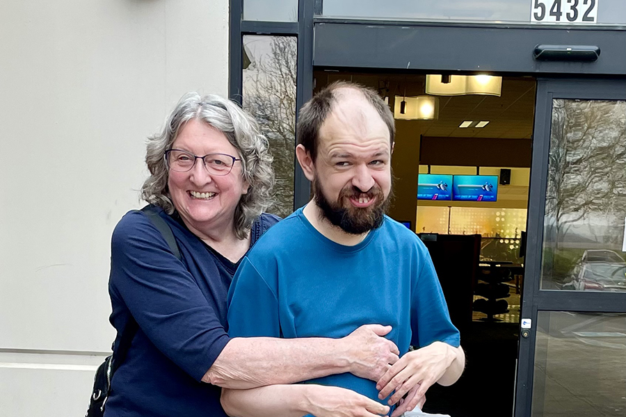 A woman stands hugging her adult son from behind.