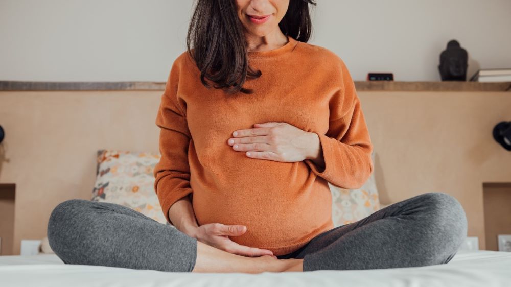 A pregnant person in an orange sweatshirt sits upright on a bed, with hands on the top and bottom of their belly.