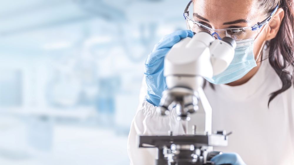 A lab researcher looks through a microscope in front of a blurred background.