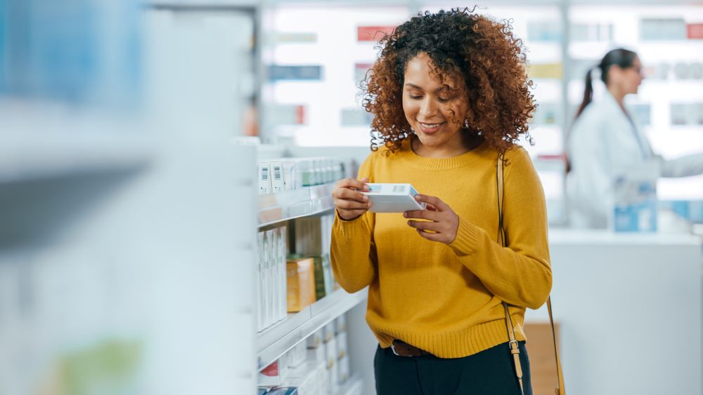 A woman a yellow shirt stands in a store pharmacy looking at a box of medicine and smiling.