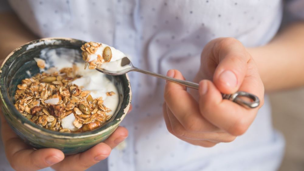 A person in a read shirt eats yogurt with muesli, nuts and pumpkin seeds.