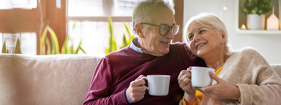 A senior couple looks at each other and has coffee while sitting closely together on a couch.