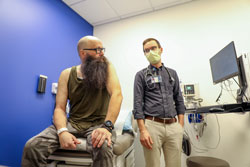 A man with cancer gets a checkup. He sists on an exam table. A physician assistant stands next to him.