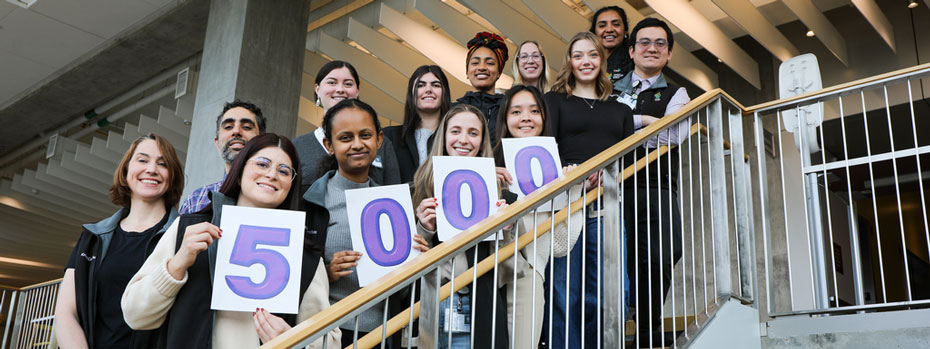  A dozen researchers stand on a staircase to celebrate a recruitment milestone. They are smiling and  holding up cards that spell out the number five thousand.