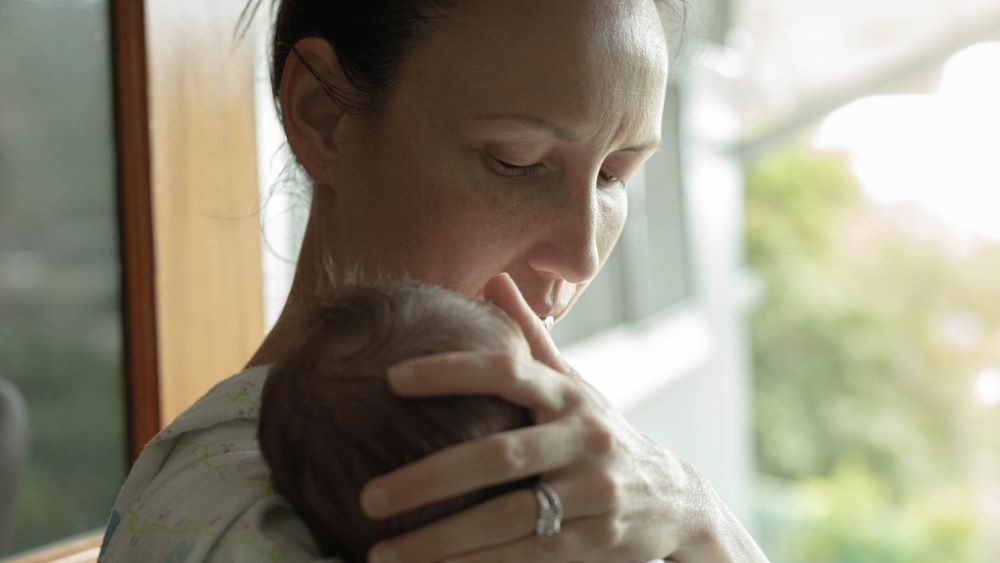 A mother stands calmly in front of a bright window with a newborn on her shoulder.