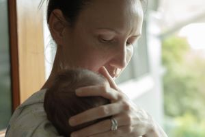 A mother holds her newborn standing in front of a door or window with the baby's head resting on her shoulder.