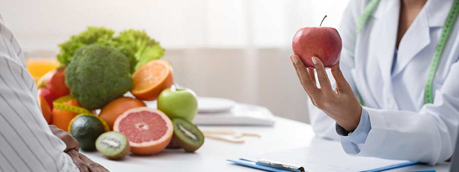 A health care provider holds an apple while sitting across from another person. A pile of fresh fruit and vegetables is between them.