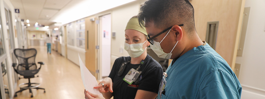 A nurse wearing dark blue scrubs points at a document, reviewing it with a trainee wearing teal scrubs. Both wear masks and stand in a hospital hallway.