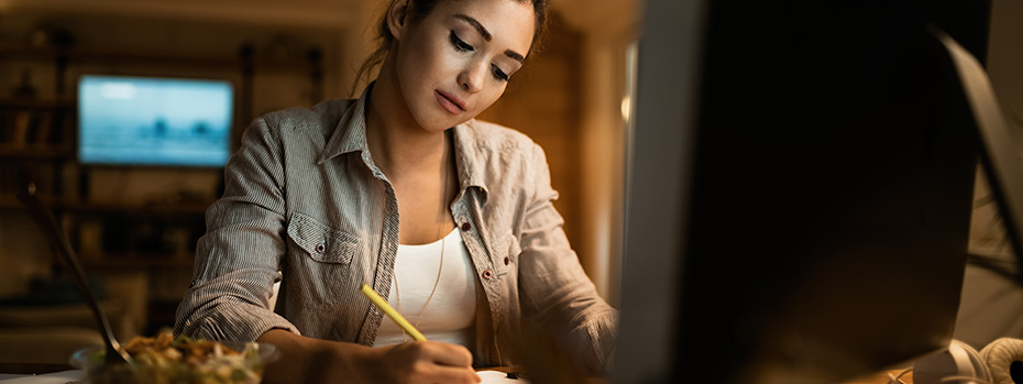 A student sits at a computer, holding a pencil doing homework. A bowl of snacks rests at her righthand side.