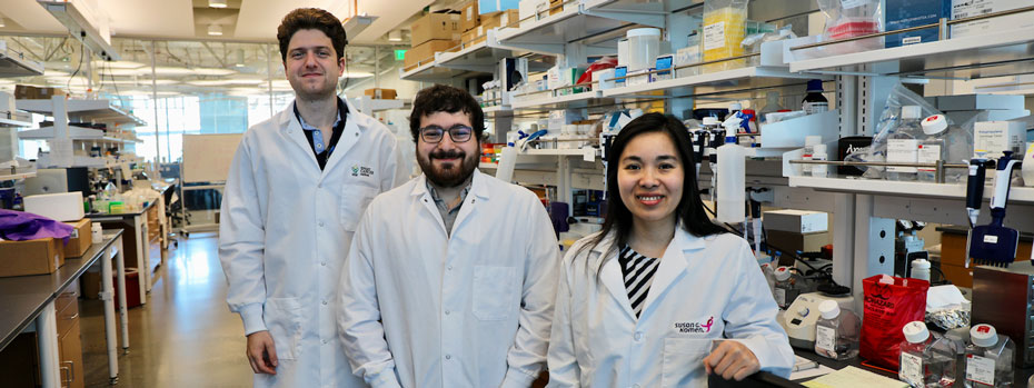 Three cancer researchers in lab coats smile as they stand at a bench filled with equipment.