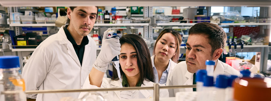 Researchers observe a teammate using a pipette in a lab filled with equipment.