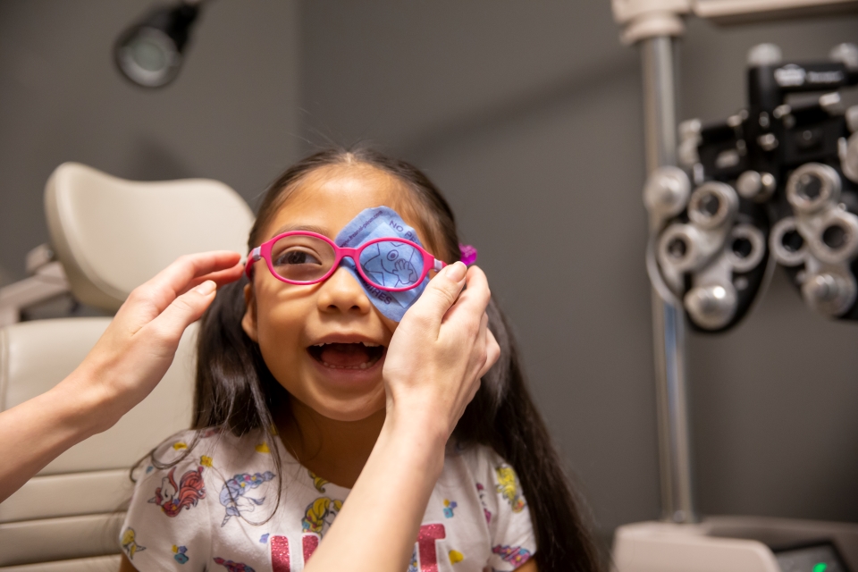 A young girl smiles at the doctor while wearing a blue patch over her left eye. A pair of hands is reaching out placing pink glasses frames on her face.