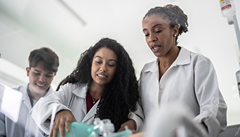 Three medical students in lab coats practice skills during a simulation.