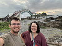 Two people posing for selfie on rocky beach in front of a bridge.