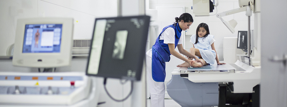 A technician adjusts a child’s leg as the child sits atop an X-ray machine.