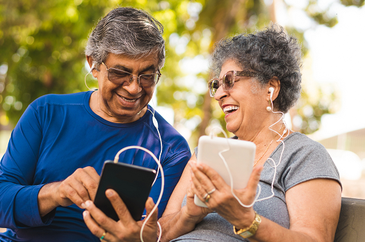 Two senior adults smiling and listening to digital devices together with earbuds outside