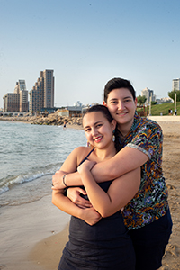 Two people hug while posing on the beach with high-rise buildings in the backgroud.