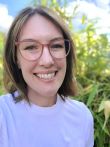 Image of a Caucasian woman with straight brown hair and glasses. She is standing in front of a blue sky and vegetation. 