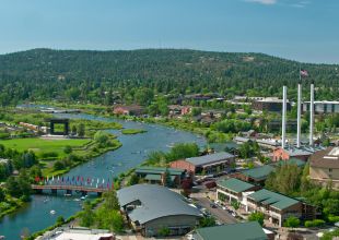 An aerial shot of the town of Bend, Oregon. It includes the Deschutes River, the Old Mill, and mountains in the background on a sunny day.