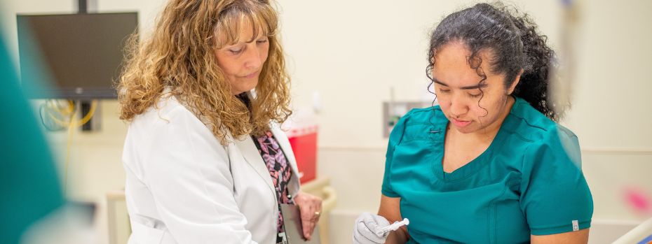 A nursing faculty member teaches to a nursing student, who is holding a needle