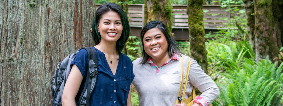 Two nursing students smile at the camera. They are standing in front of a large tree trunk and have ferns and other plants behind them.