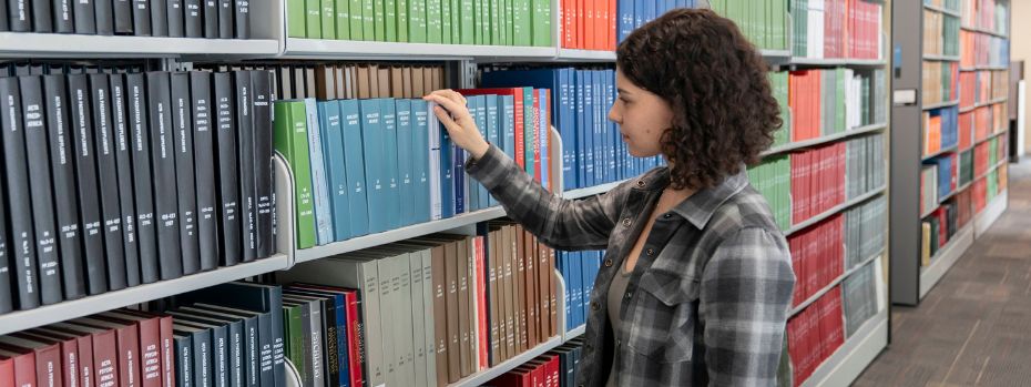 A student in a library chooses a blue book off the shelf to read.