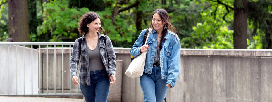 Two nursing students chat with each other as they walk towards the camera. There's a short wall behind them and trees.