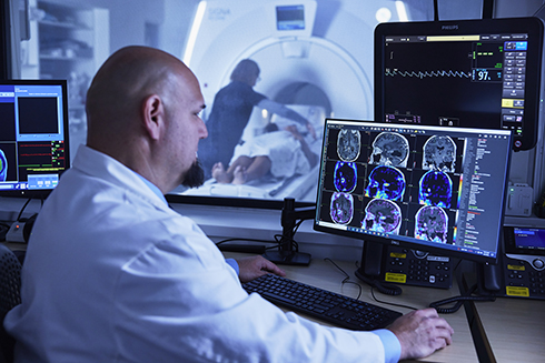 OHSU neuroradiologist Ramon Barajas looks at PET/MRI images on a computer monitor. In the next room, visible through a window, a technologist helps a person lying on the bed of the PET/MRI scanner.