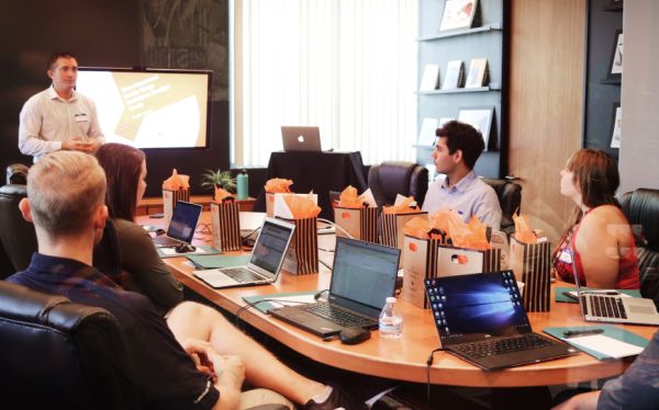 A man gives a presentation to a group of people at a conference table who are taking notes on laptop and looing attentively towards the presentation.