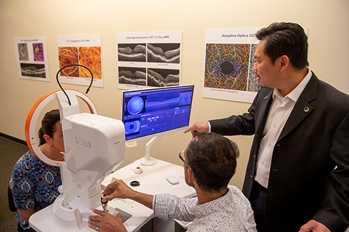 Dr. David Huang stands behind a technician looking at a screen with images of the eye of a person seated at a retina scanner.