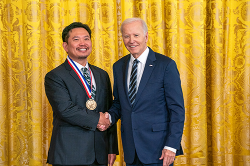 Dr. David Huang wears the National Medal of Technology and Innovation on a red, white and blue ribbon around his neck. He is shaking hands with President Joe Biden in front of a yellow curtain.