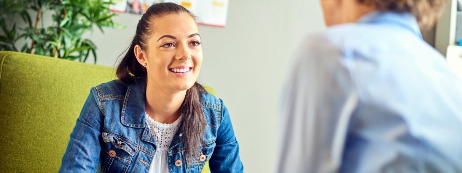 A young woman sits facing the camera while she speaks with an another person, whom we can only see from the back.