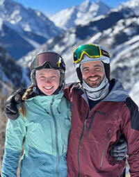 A man standing with his arm around a woman, both smiling, while on a mountain skiing or snowboarding.