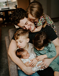 A man sitting in a chair with two toddlers in his lap, one of whom is holding a baby, and a third toddler hanging on his back.