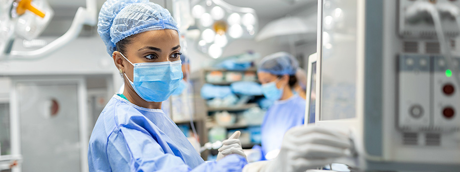 An anesthesia nurse practitioner wearing scrubs and face mask adjust equipment in an operating room. 