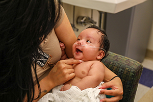 A baby with a breathing tube taped to his face smiles at his mother as she holds him on her lap.