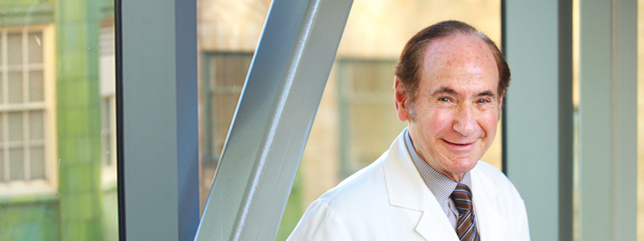 Photo of Dr. Albert Starr in his white physician coat leaning against a glass wall overlooking the OHSU campus.