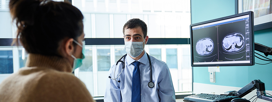 OHSU cancer specialist Guillaume Pegna sits with a person in an exam room. A large computer monitor showing imaging scans is next to him.