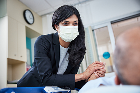 OHSU cancer surgeon Divya Sood leans toward a person lying in a hospital bed and clasps one of their hands in both of hers.