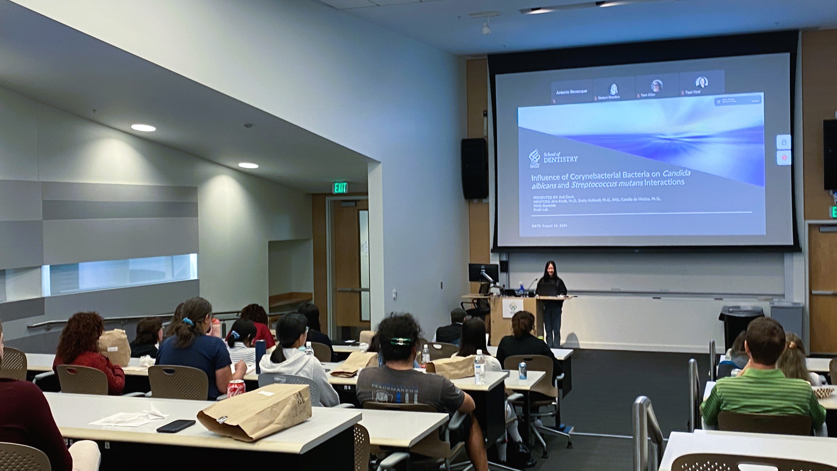 Cover slide of a presentation shows on the large screen of a classroom, as a seated crowd gives their attention to a person standing at a lectern underneath the large screen.