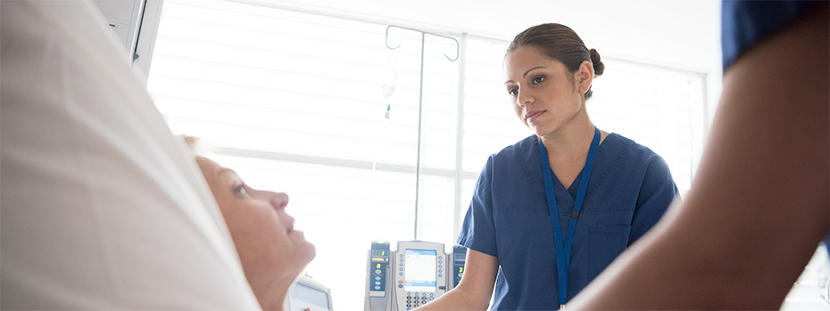 An OHSU nursing student in blue scrubs stands at a patient’s bedside, listening attentively.