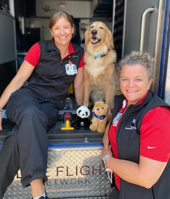 A member of the pediatric and neonatal transport team at OHSU Doernbecher Children’s Hospital sits in the back of an ambulance with Davis the golden retriever. Another team member stands next to the ambulance. A toy panda and toy golden retriever sit in front of Davis.