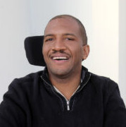 A parson with dark tan skin and dark short hair wearing a black quarter-zip sweater and sitting in a power wheelchair.