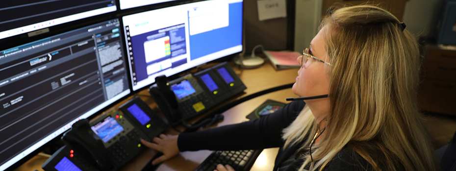 A person wearing a headset presses a button on a phone in the middle of a row of phones. The person is sitting before a bank of monitors showing data. 