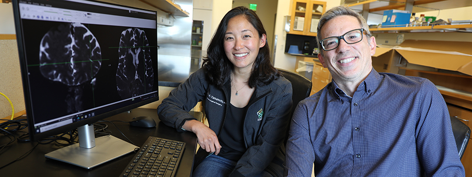 Dr. Erin Yamamoto and Dr. Juan Piantino sit next to a computer screen that shows brain scan images.