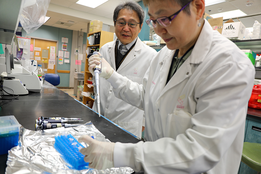 A researcher in a lab holds a liquid dropper over test tubes as another researcher looks on.