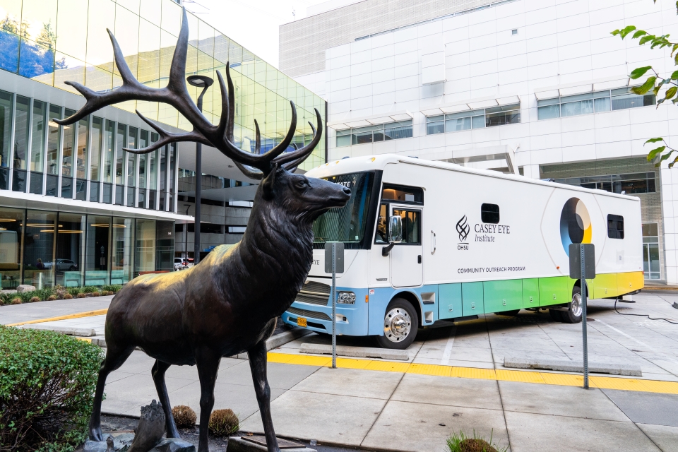 A statue of an elk sits in the foreground, with the Casey Community Outreach Program mobile clinic in the center, and the Casey Eye Institute and the Elks Children's Eye Clinic buildings in the background.