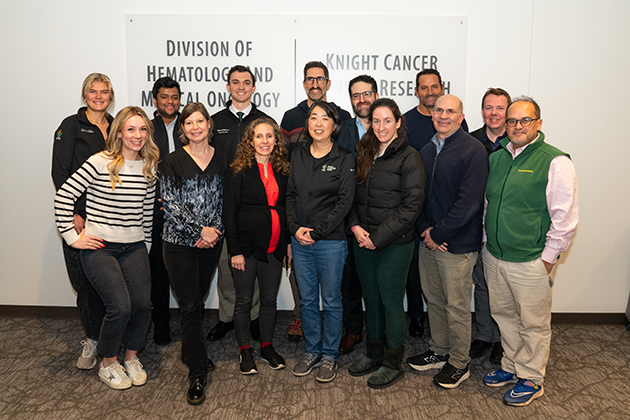 A group photo of the GI cancer research group. There are 14 men and women standing in front of their office space at OHSU. 