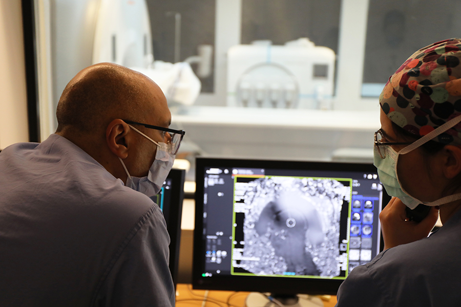 Two health care providers wearing PPE facemasks sitting at a desk and looking at a monitor displaying a medical scan.